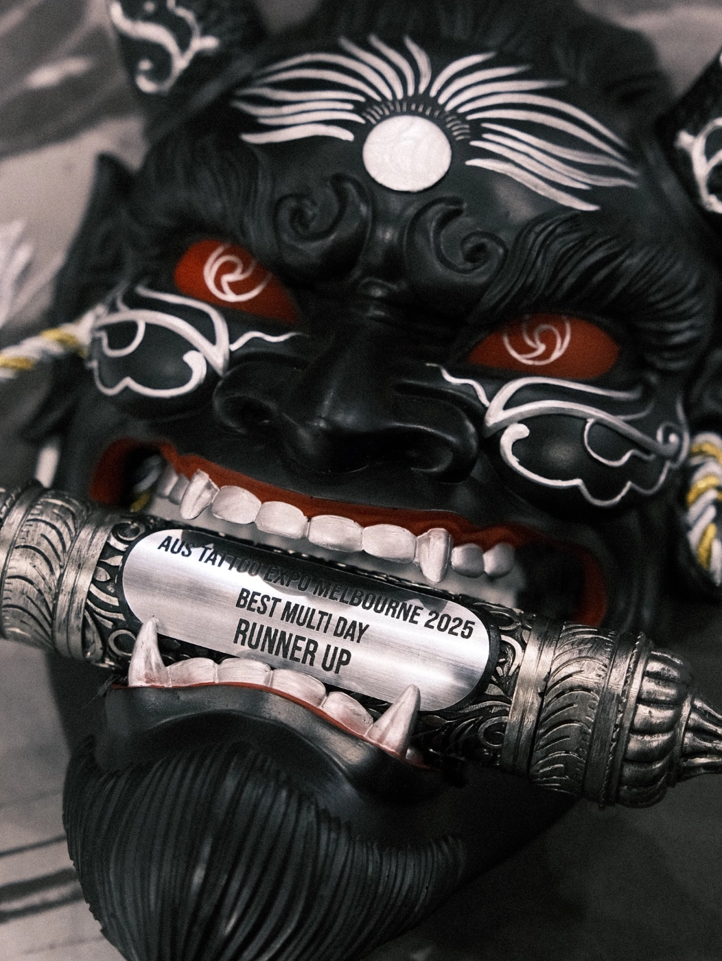 Illustrative blackwork tattoo of a demon mask with an award scroll.