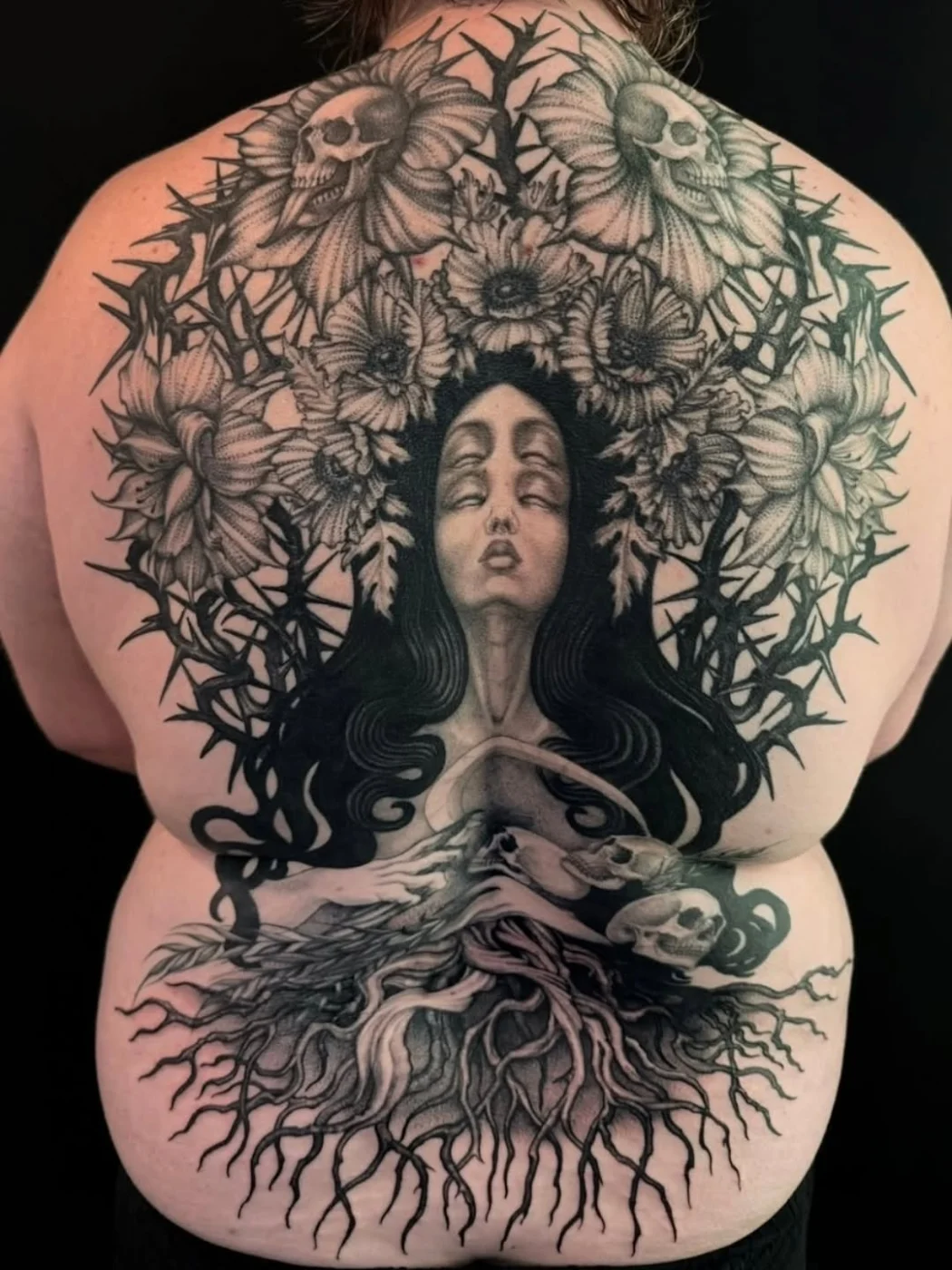 Large black-and-grey tattoo of a woman with flowers and skulls on back.