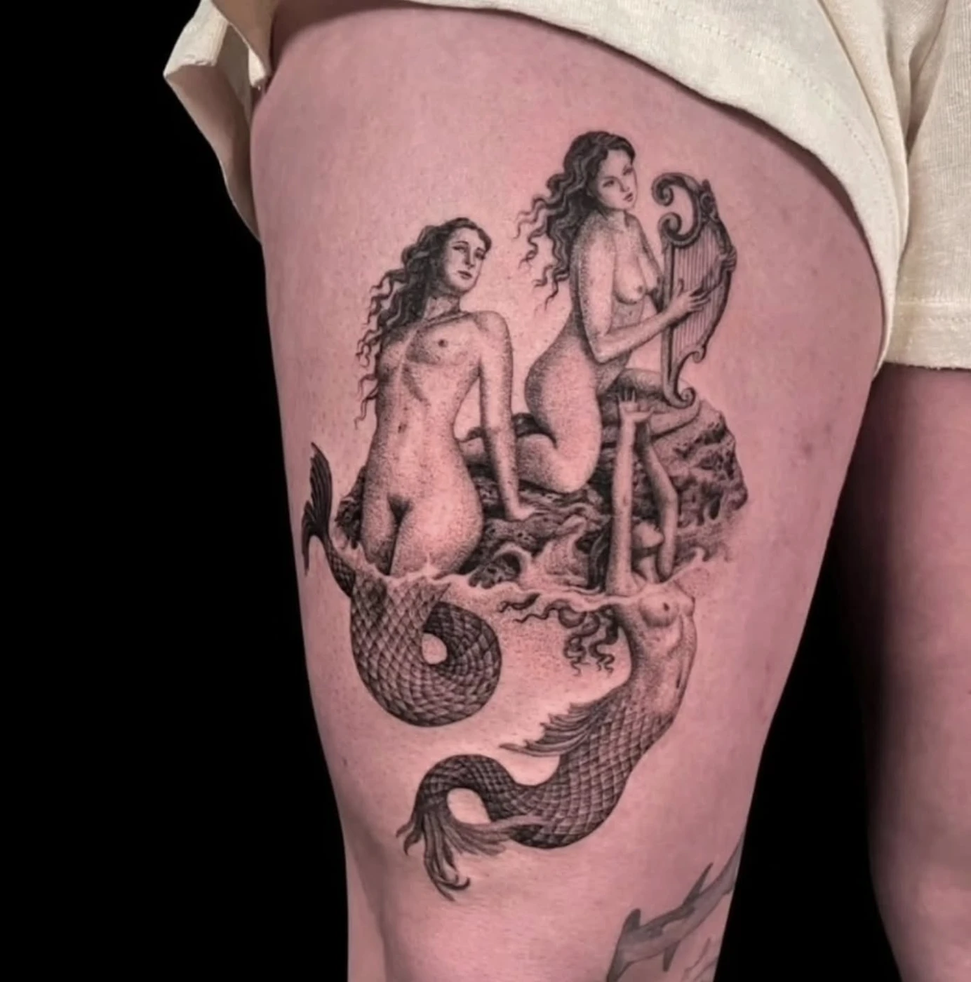 Black-and-grey mermaid tattoo on thigh featuring two female figures.
