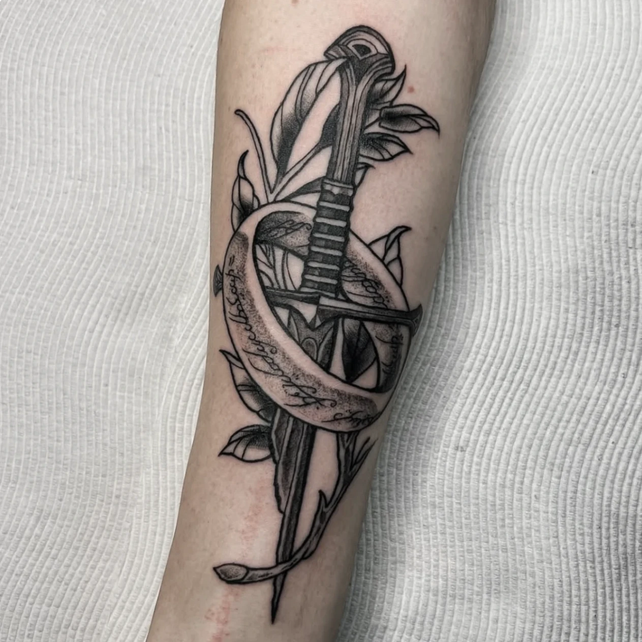 Black-and-grey sword tattoo with snake and leaves on forearm.