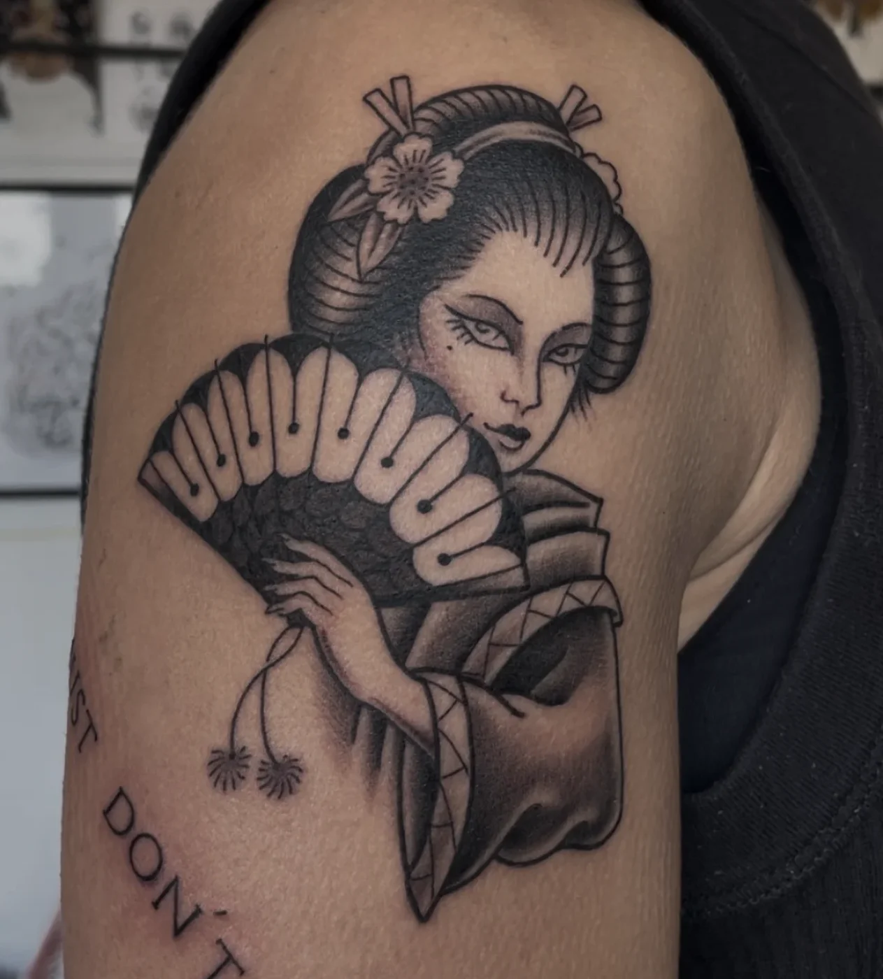 Black-and-grey tattoo of a geisha holding a fan on shoulder.